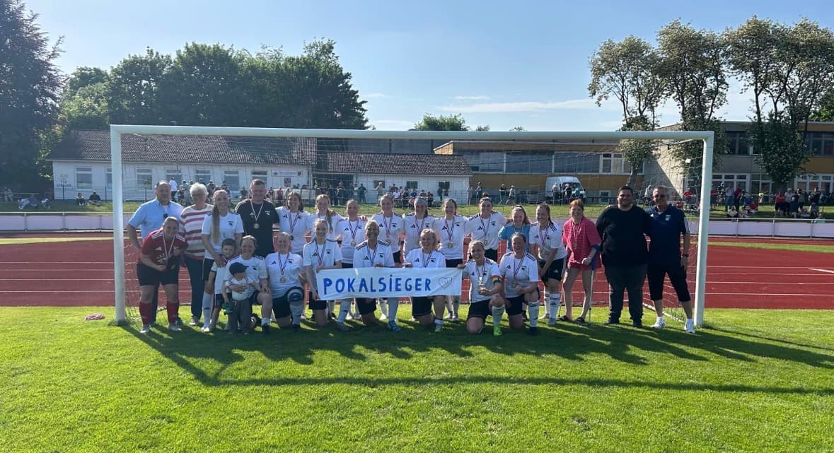 Die Fußball-Damen mit Medaillen und Pokalsieger-Banner vor dem Tor auf dem Sportplatz