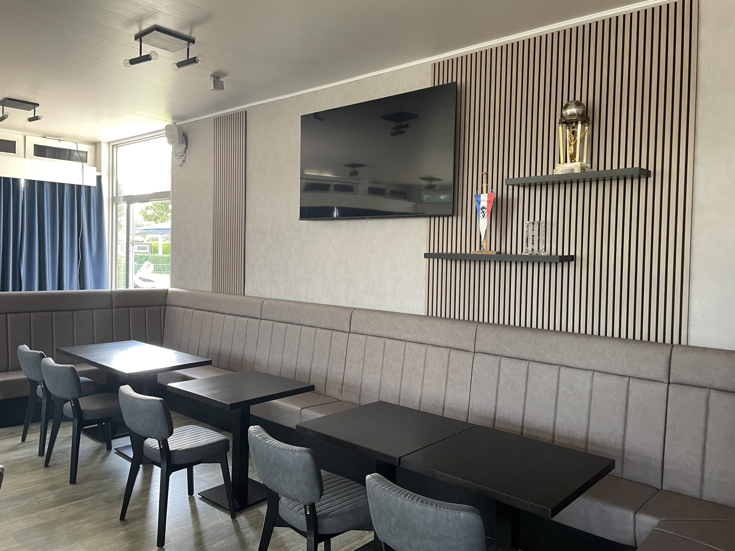 Ein modernes, leeres Café mit grau gepolsterten Sitzmöbeln, schwarzen Tischen und Stühlen, einem großen, an der Wand montierten Fernseher und zwei Trophäenauslagen auf einem Regal über dem Sitzbereich. Natürliches Licht fällt durch ein Fenster mit dunklen Vorhängen ein.
