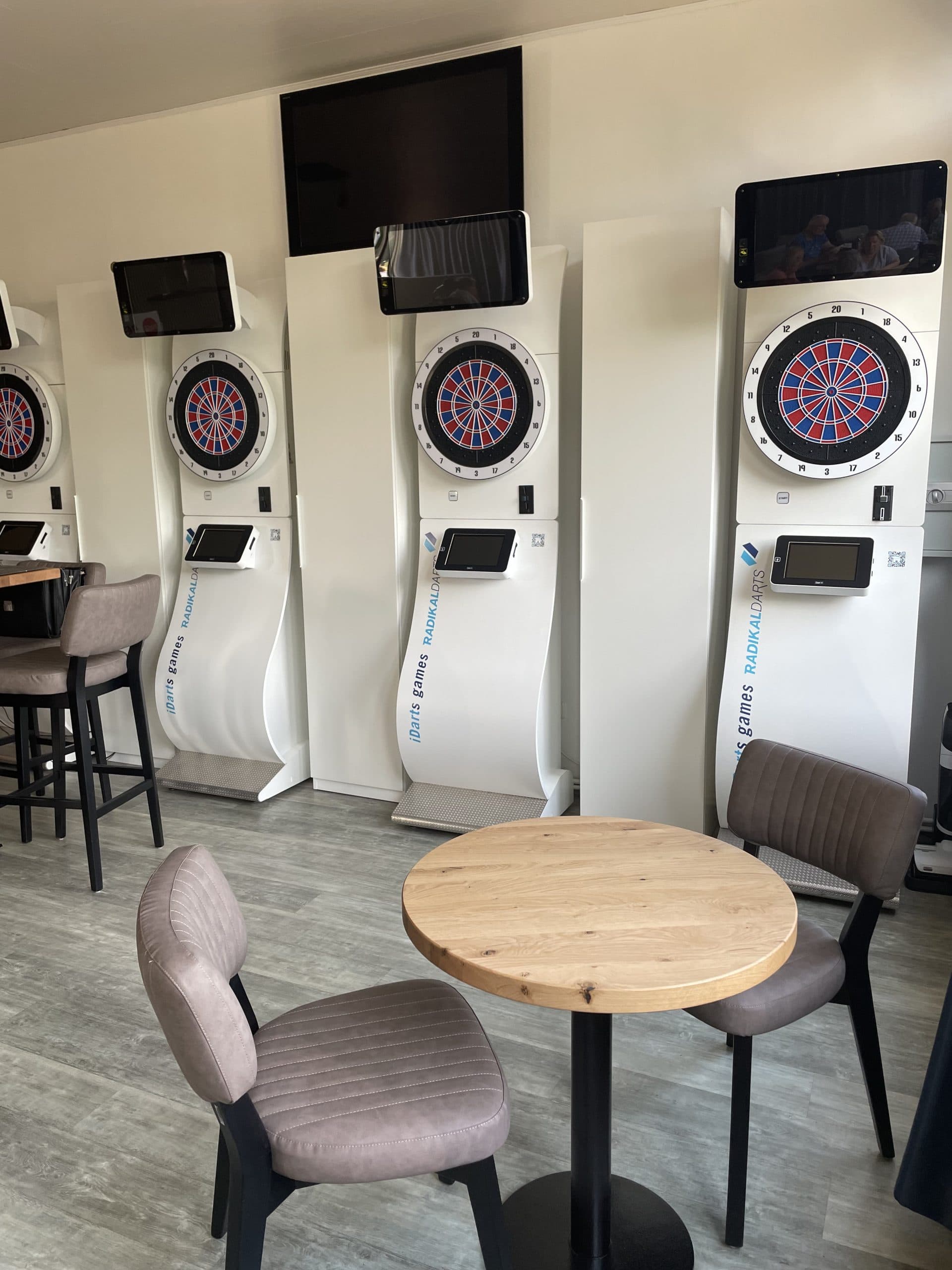 Drei elektronische Dartscheiben sind an einer Wand in einem modernen Raum mit hellem Holzfußboden angebracht. Im Vordergrund stehen ein runder Holztisch und zwei gepolsterte Stühle, links davon sind Stehtische und Stühle zu sehen.