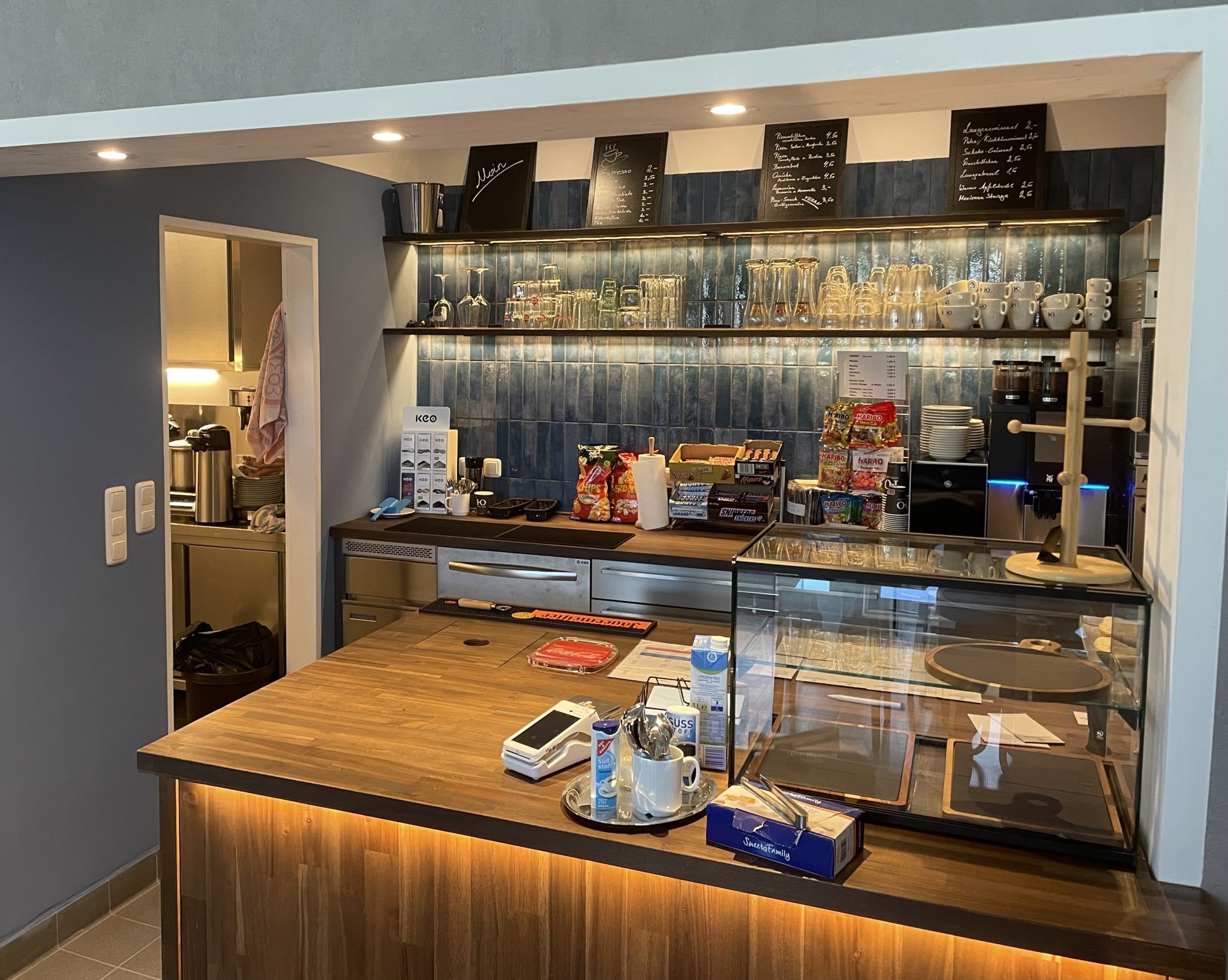 Eine gemütliche Café-Theke mit einer hölzernen Arbeitsplatte, einer Glasvitrine, einer Kaffeemaschine, Snacks und Regalen mit Tassen und Gläsern. Im Hintergrund ist ein Küchenbereich zu sehen. Die Speisekarten sind auf Tafeln darüber geschrieben.