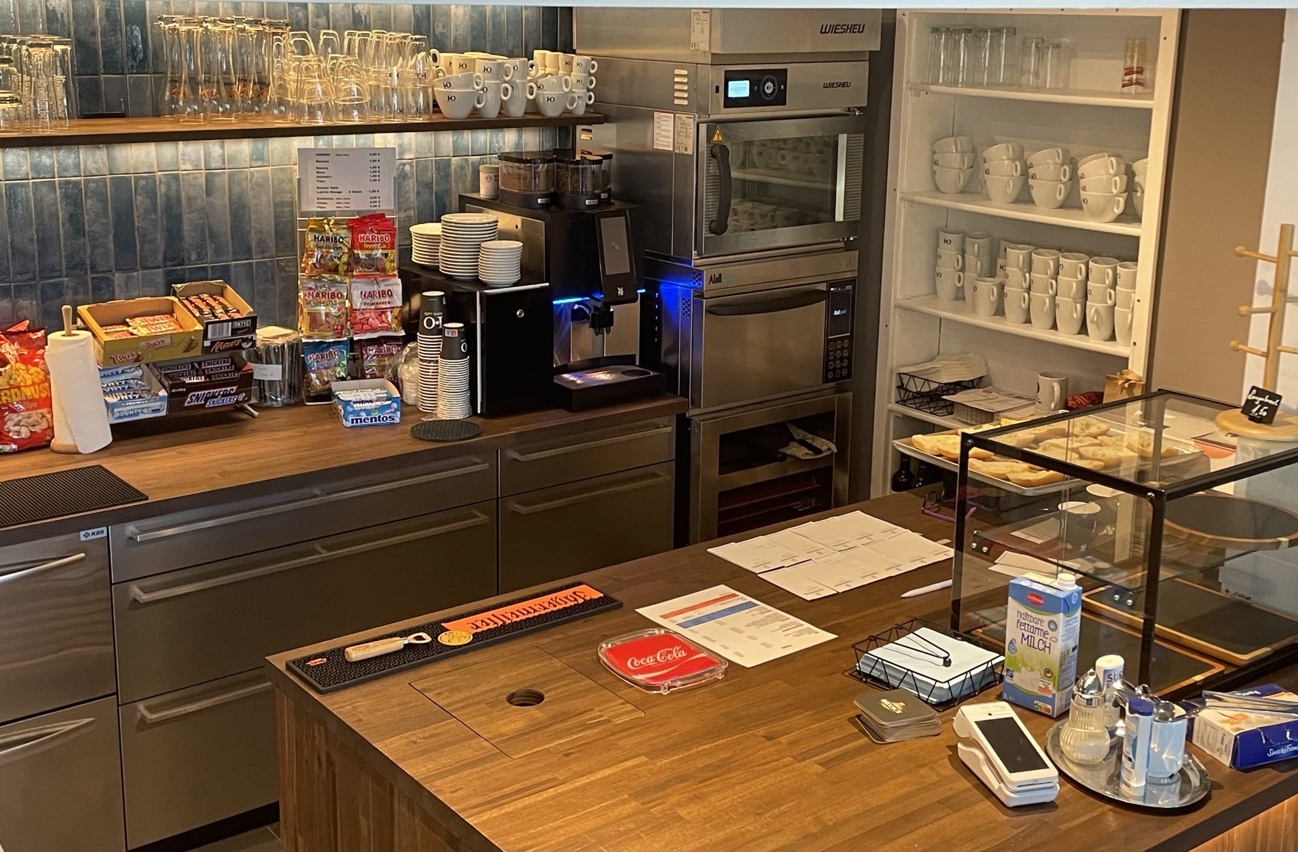 Eine Kaffeetheke mit einer Kaffeemaschine, einer Mikrowelle, Snacks, Tassen, einer Glasvitrine mit Gebäck und verschiedenen Gegenständen wie Speisekarten und Gewürzen auf der hölzernen Arbeitsplatte. Im Hintergrund befinden sich Regale mit Gläsern und Tassen.