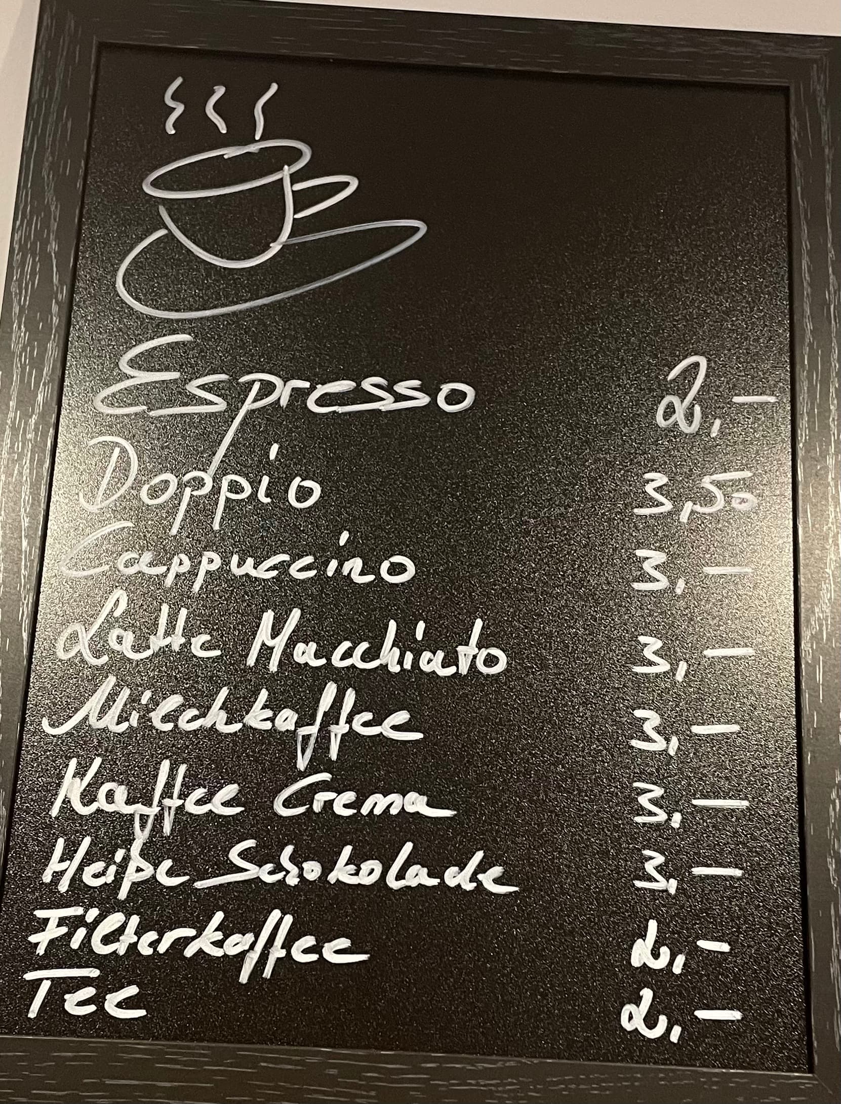 Auf einer Tafel mit einer illustrierten Kaffeetasse sind die Getränke auf Deutsch aufgelistet: Espresso, Doppio, Cappuccino, Latte Macchiato, Milchkaffee, Kaffee Crema, Heiße Schokolade, Filterkaffee und Tee, mit Preisen von 2€ bis 3,50€.