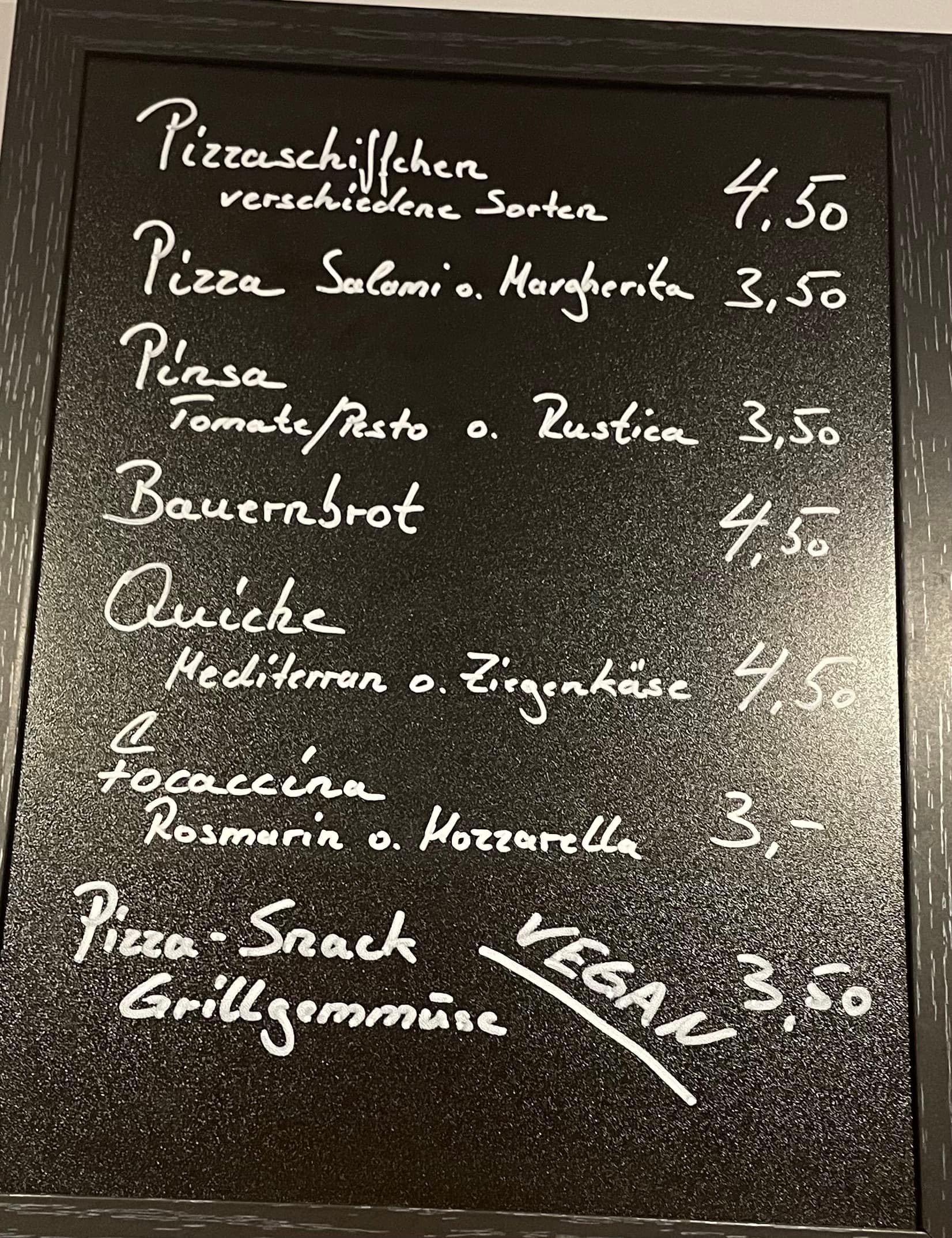 Eine Speisekarte mit schwarzer Kreidetafel und weißer Handschrift mit deutschen Speisen und Preisen, darunter Pizza, Pina, Quiche, Focaccia und ein veganer Pizzasnack. Die Preise reichen von 3 bis 4,50 Euro.