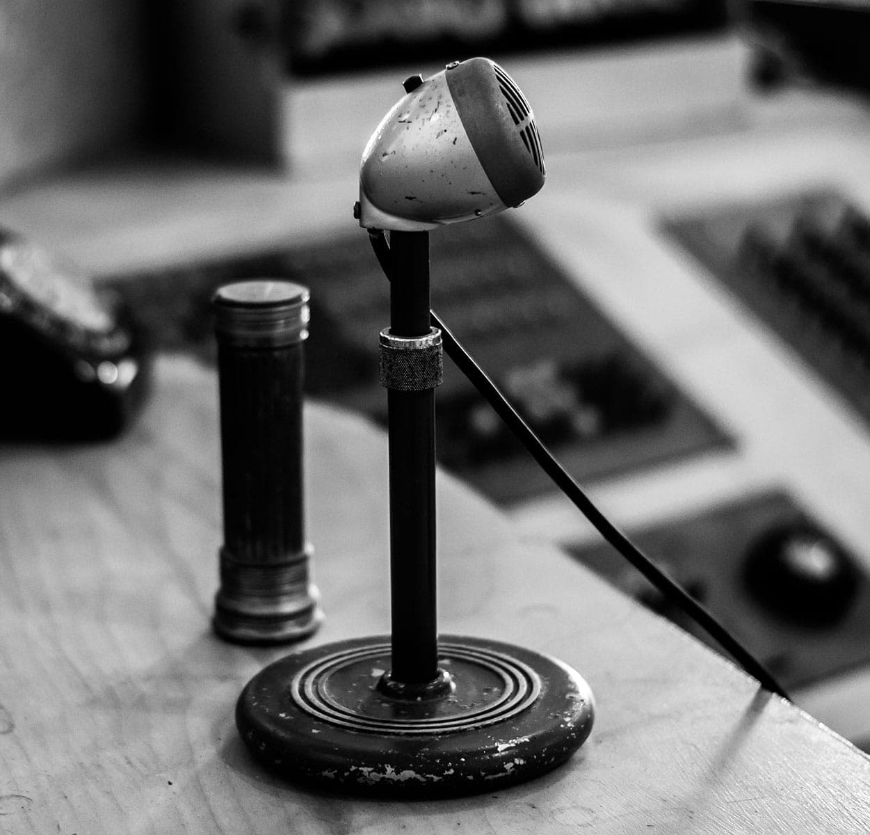 Ein Schwarz-Weiß-Foto eines alten Mikrofons auf einem runden Sockel, an dem ein Kabel befestigt ist, das auf einem Schreibtisch neben einem zylindrischen Objekt steht. Im Hintergrund sind unscharfe elektronische Geräte zu sehen.