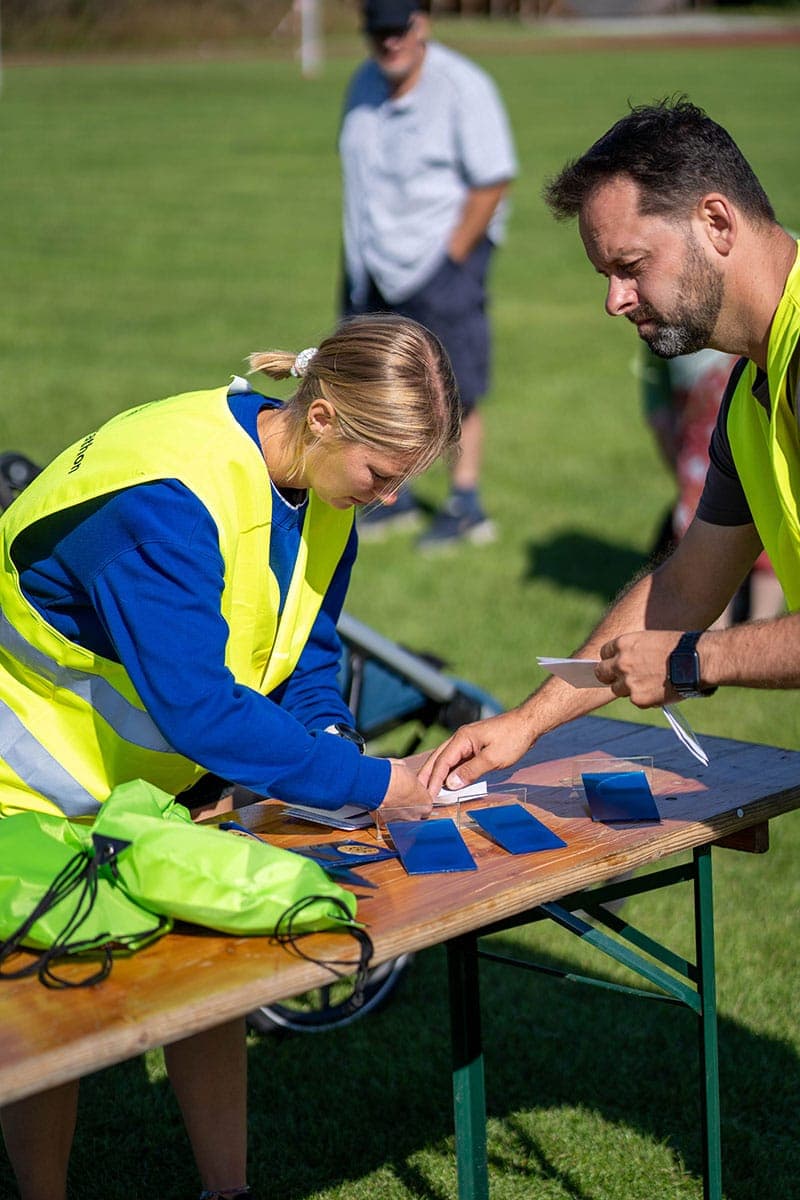 Zwei Personen in gelben Sicherheitswesten arbeiten an einem Tisch im Freien und sortieren blaue Karten, während auf dem Tisch grüne Taschen liegen und im Hintergrund eine Wiese zu sehen ist. Eine weitere Person steht in der Ferne.