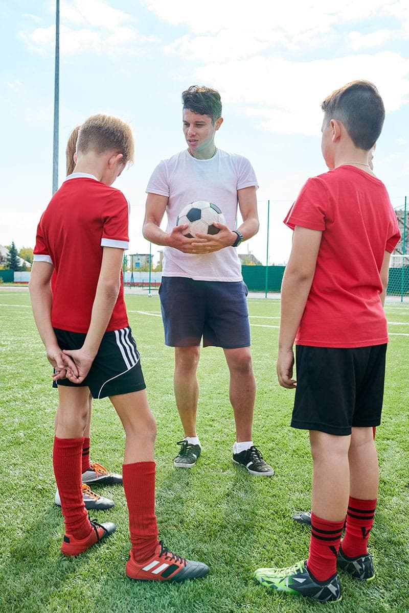 Ein Fußballtrainer, der einen Ball in der Hand hält, spricht mit drei Jungen in roten Trikots, die auf einem grünen Fußballfeld stehen, wobei zwei Jungen wegschauen und einer nach unten blickt.