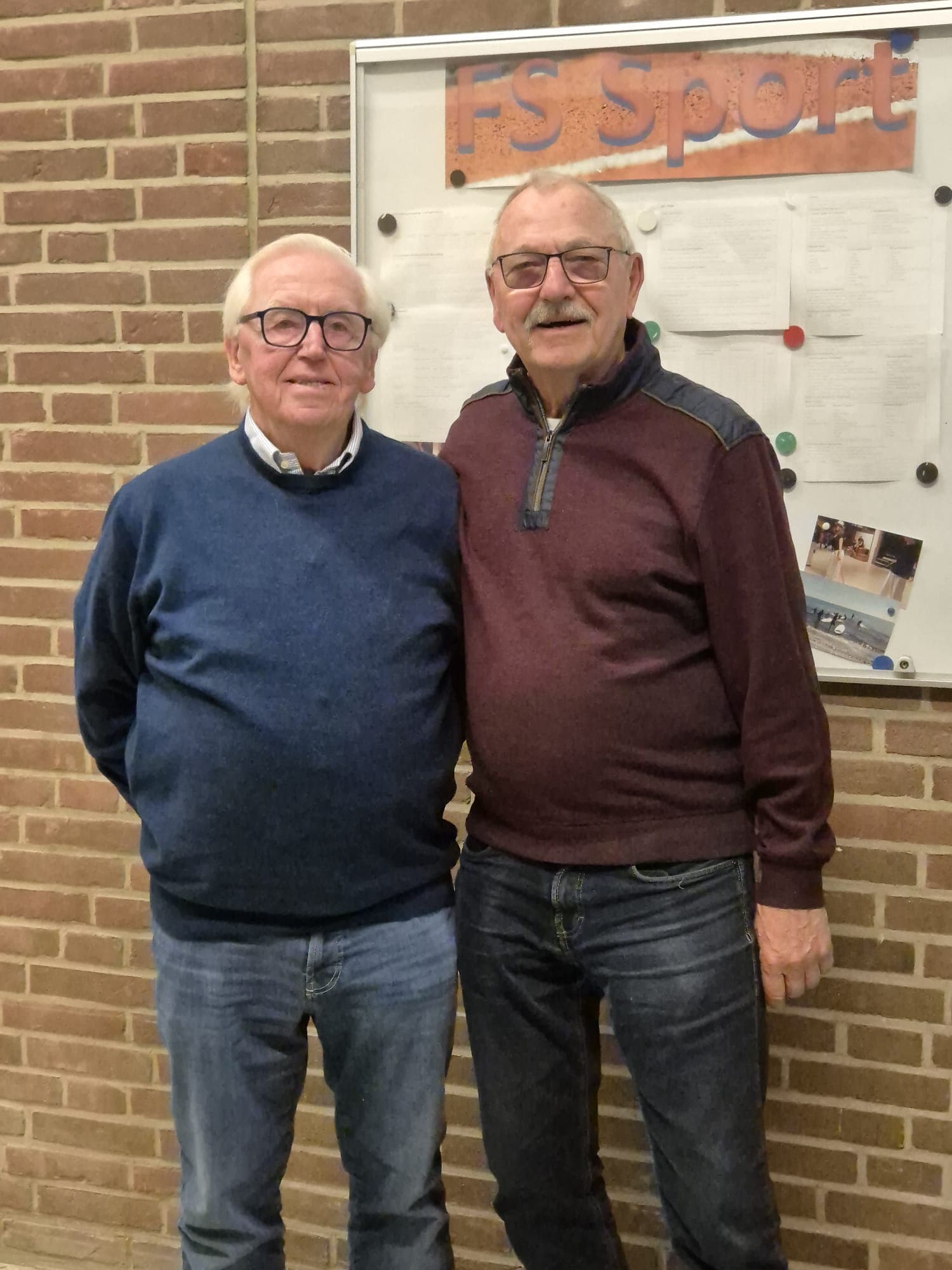 Zwei ältere Männer stehen lächelnd nebeneinander vor einer Backsteinwand und einer Pinnwand mit der Aufschrift FS Sport. Beide tragen Brillen und Pullover, sie wirken entspannt und freundlich.