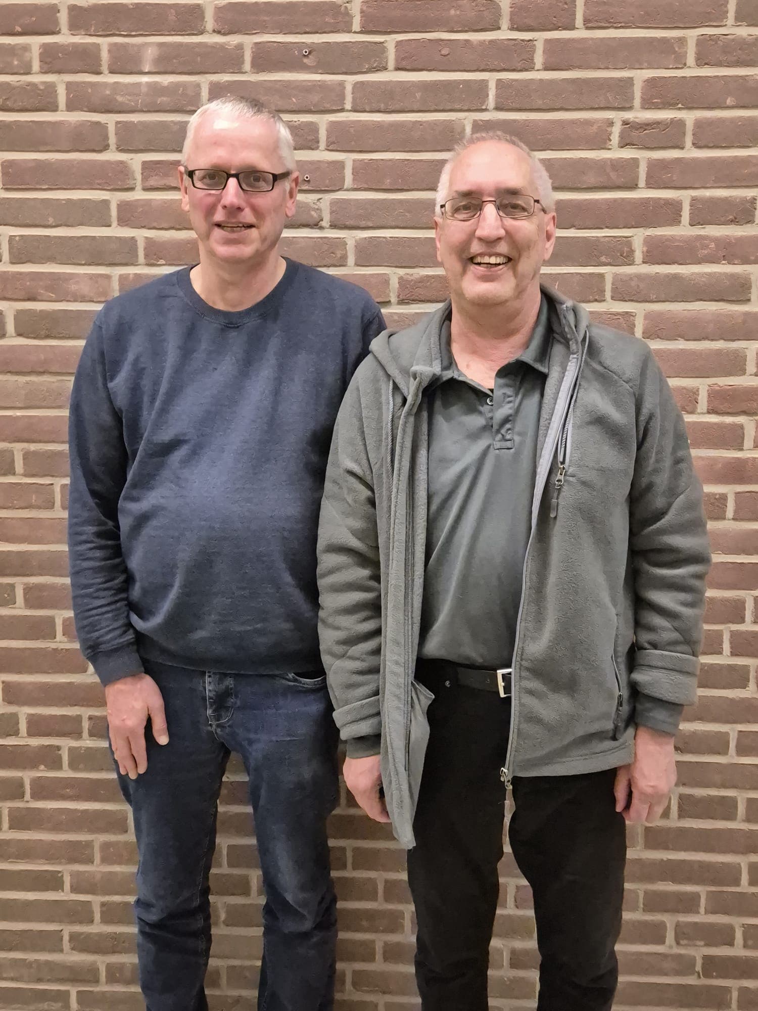 Zwei ältere Männer mit Brillen stehen lächelnd nebeneinander vor einer Backsteinmauer. Der eine trägt einen blauen Pullover und Jeans, der andere eine graue Jacke über einem Poloshirt und eine schwarze Hose.