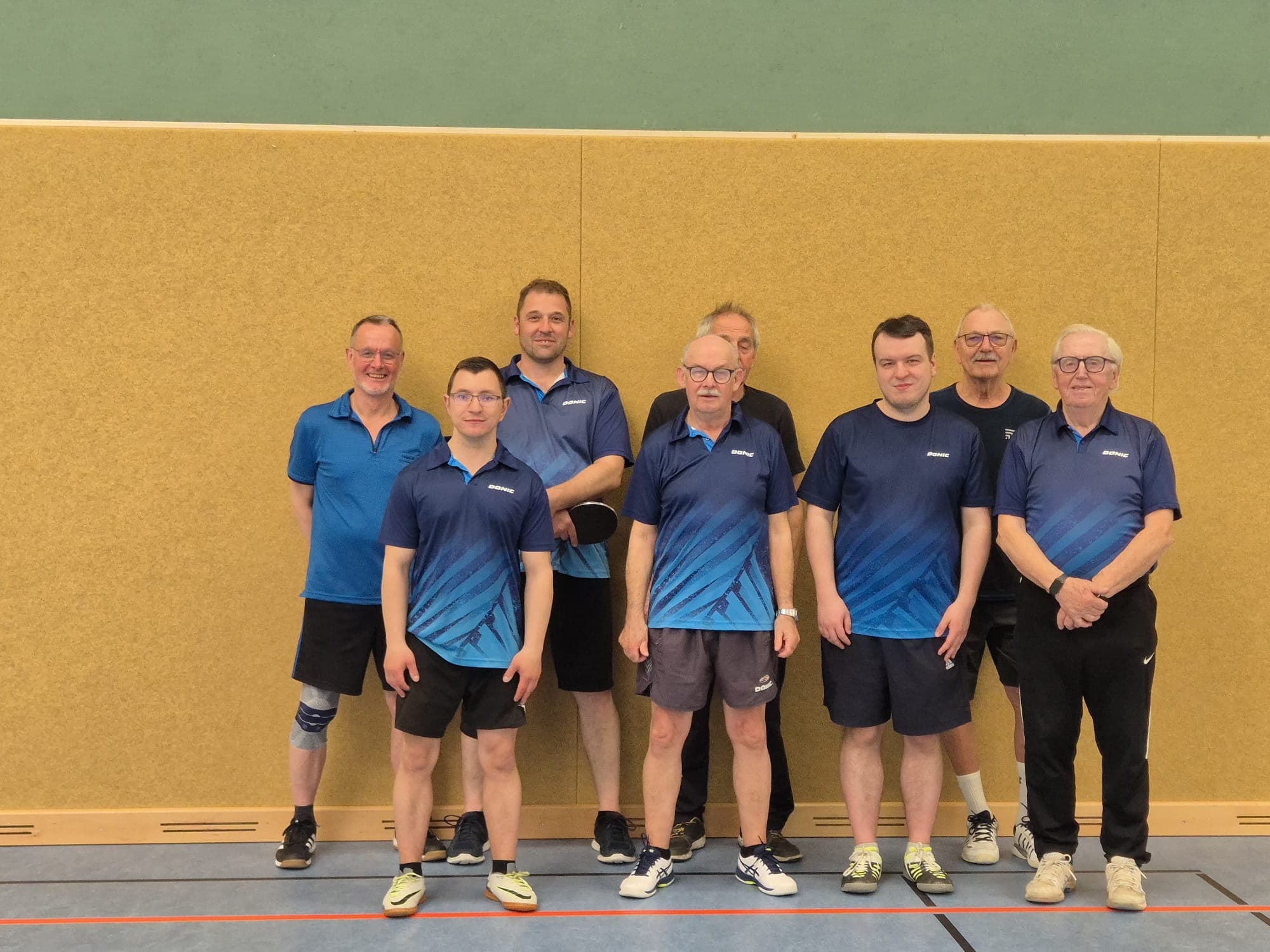 Acht Männer in Sportkleidung stehen in zwei Reihen in einer Turnhalle, hinter der eine Holzwand steht. Die meisten von ihnen tragen passende blaue Hemden, und sie scheinen alle bereit für ein Sportereignis oder ein Mannschaftsfoto.