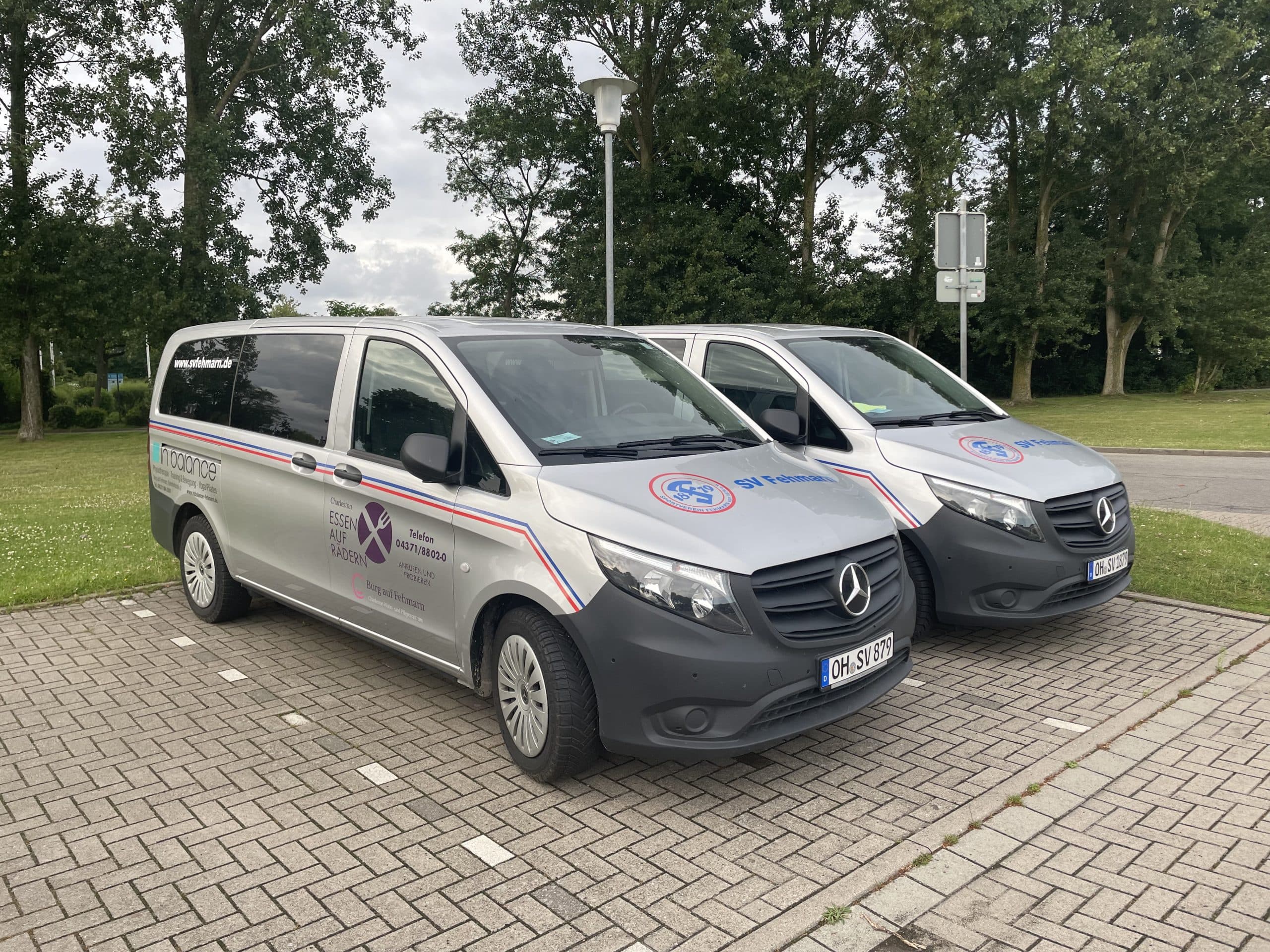 Zwei silberne Mercedes-Benz-Kleintransporter mit Firmenlogos und dem Schriftzug "Insurance" an der Seite sind auf einer gepflasterten Fläche mit grünen Bäumen und Rasen im Hintergrund geparkt.