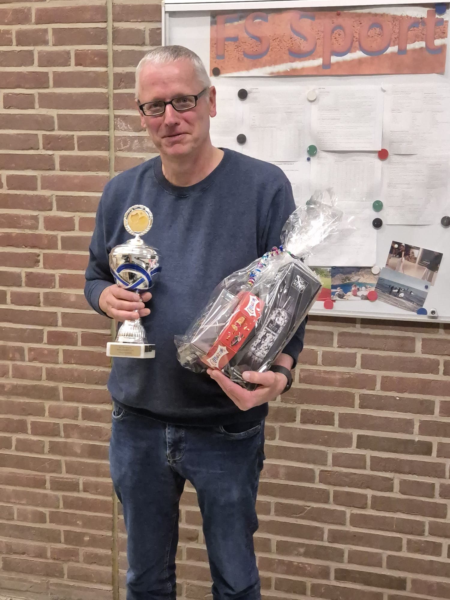 Ein Mann mit Brille, blauem Pullover und Jeans steht in einem Haus und hält in der einen Hand eine Trophäe und in der anderen einen verpackten Geschenkkorb. Im Hintergrund sind eine Anschlagtafel und eine Backsteinmauer zu sehen.