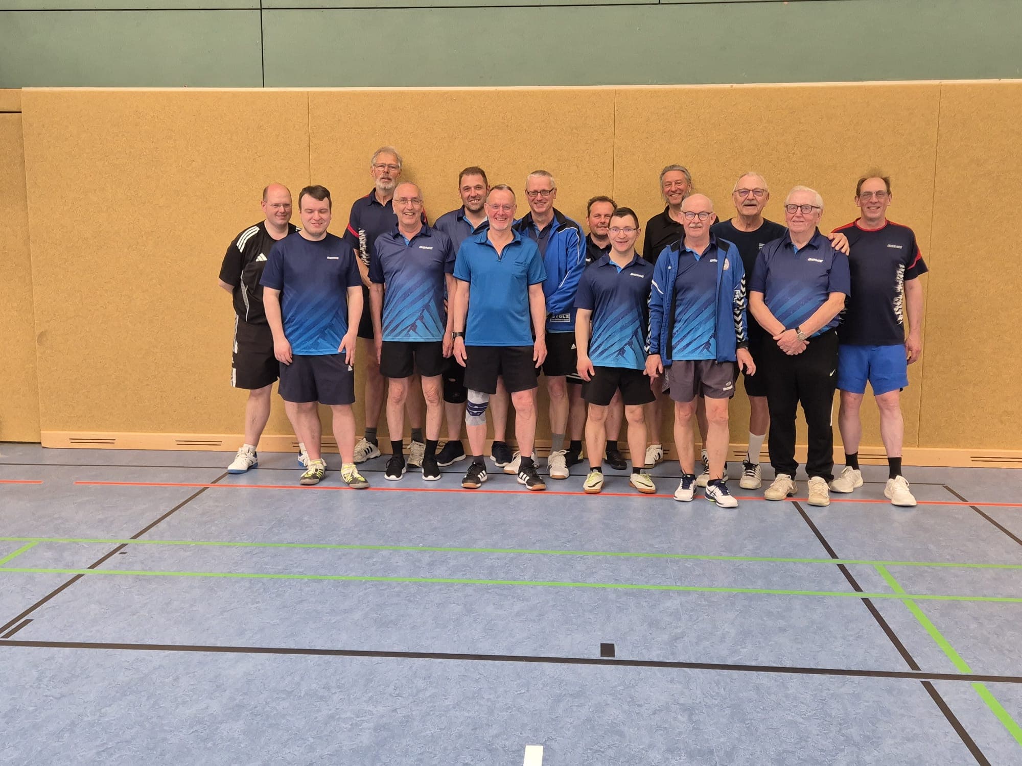 Eine Gruppe von 14 Männern, die meisten in blauer Sportkleidung, steht für ein Gruppenfoto in einer Turnhalle mit einer Holzwand und blauen Bodenmarkierungen hinter ihnen und lächelt.